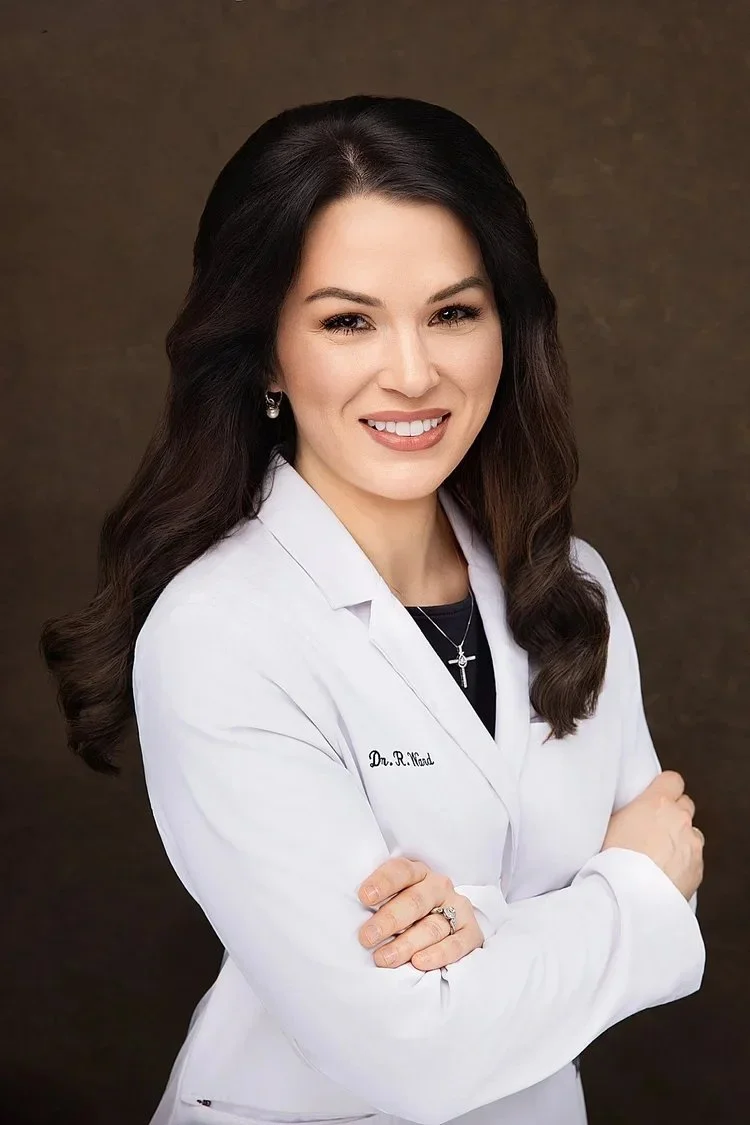 Professional portrait of a woman doctor with dark hair, wearing a white lab coat, standing with arms crossed, smiling, against a brown background.