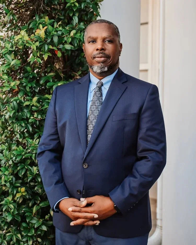 A middle-aged African American man in a navy blue suit, light blue shirt, and patterned tie standing outdoors beside a leafy green bush, with a neutral expression.