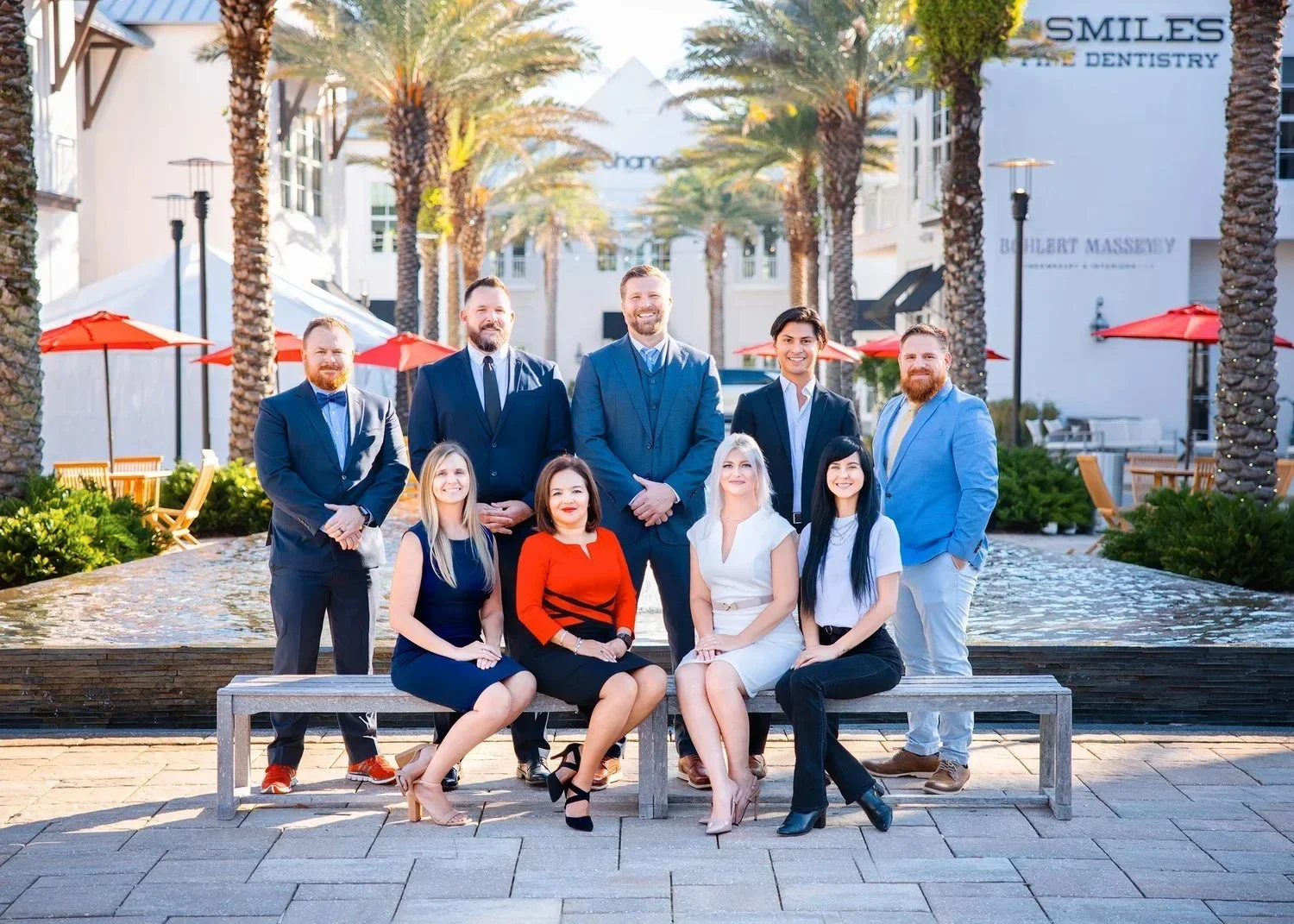Group of professionals dressed in business attire posing outdoors in front of a water feature and palm trees, with a shopping center or office complex in the background.