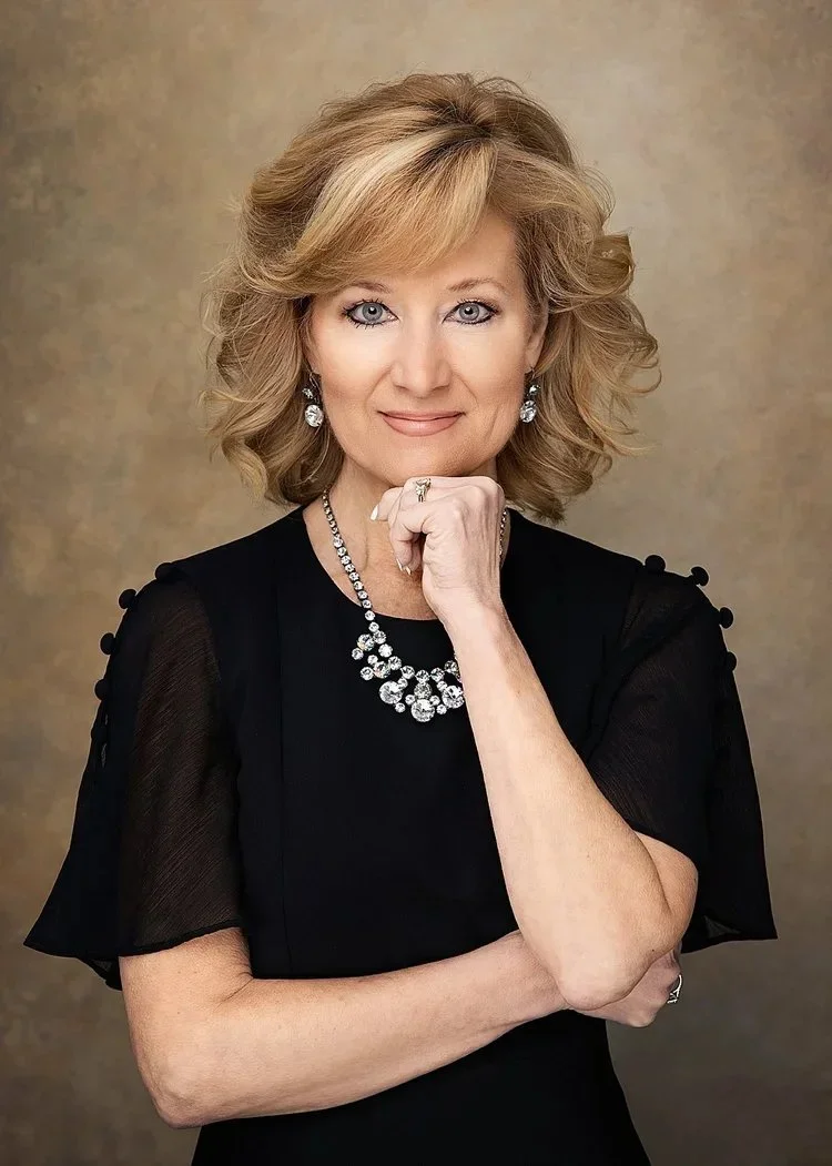 A middle-aged woman with blonde hair styled in loose waves, wearing a black dress with sheer sleeves, elegant jewelry including a necklace, earrings, and a ring, posing with her hand near her chin against a neutral background.