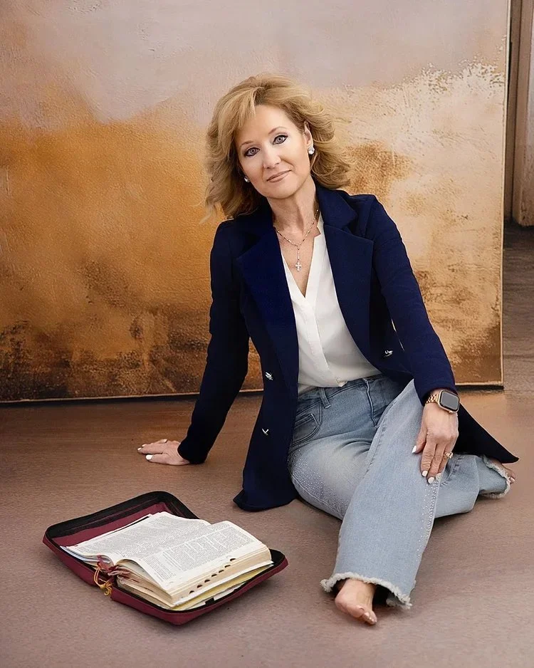A woman sitting on the floor with an open Bible or book in a case in front of her. She has blonde hair, is wearing a dark blue blazer, light gray ripped jeans, and a white blouse. She has a cross necklace and a smartwatch on her left wrist, and is po