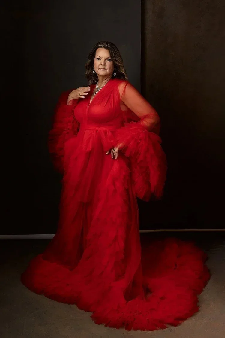 A woman wearing a red gown with sheer long sleeves and ruffled tulle details, standing against a dark background.