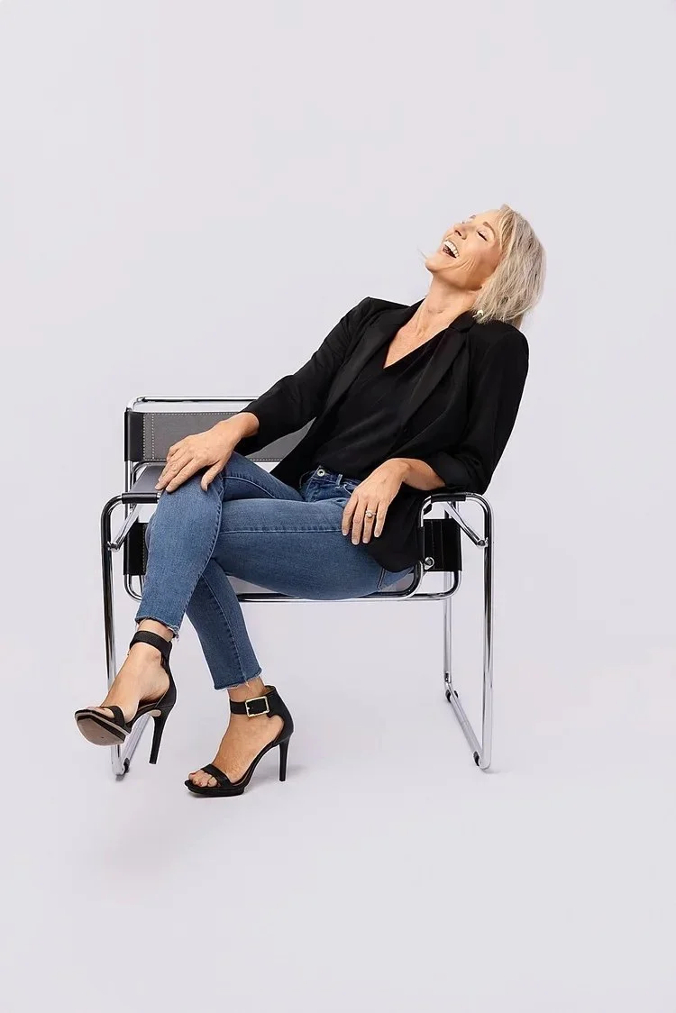 A woman with blonde hair sitting on a modern metal chair, laughing and looking upward, wearing a black blazer, black top, blue jeans, and black high-heeled sandals, against a plain white background.