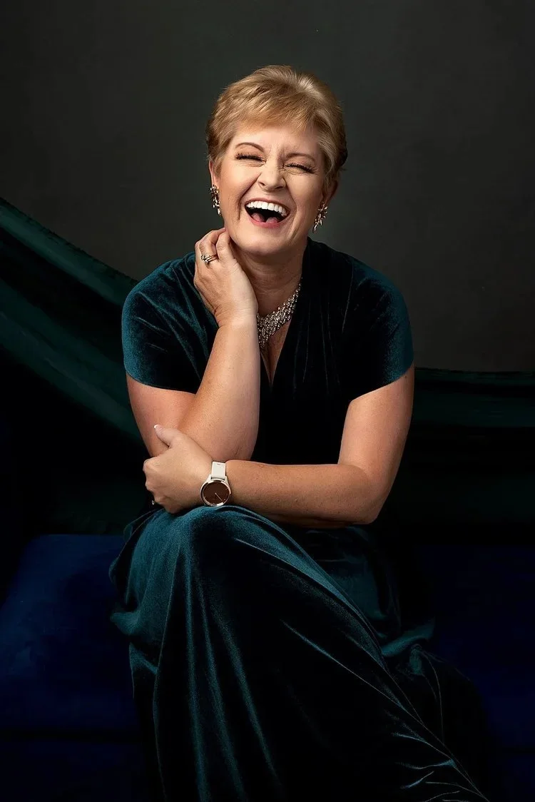 A woman with short blonde hair smiling and laughing, sitting with her hand on her chin, wearing a dark velvet dress, jewelry, and a watch, against a dark background.