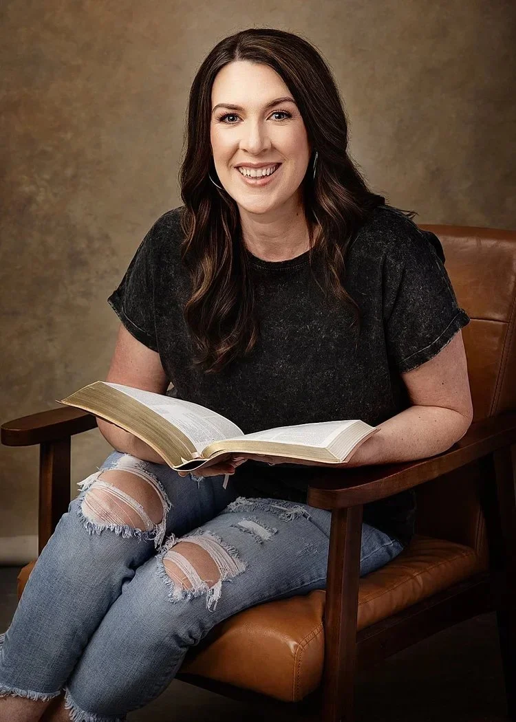 A young woman with long, dark hair, wearing a black t-shirt and ripped jeans, sitting in a brown chair, holding an open book, smiling.