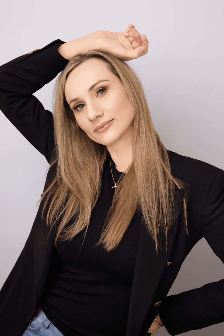 A woman with long blonde hair wearing a black blazer, black top, and a cross necklace, posing with her right hand raised above her head and smiling softly.