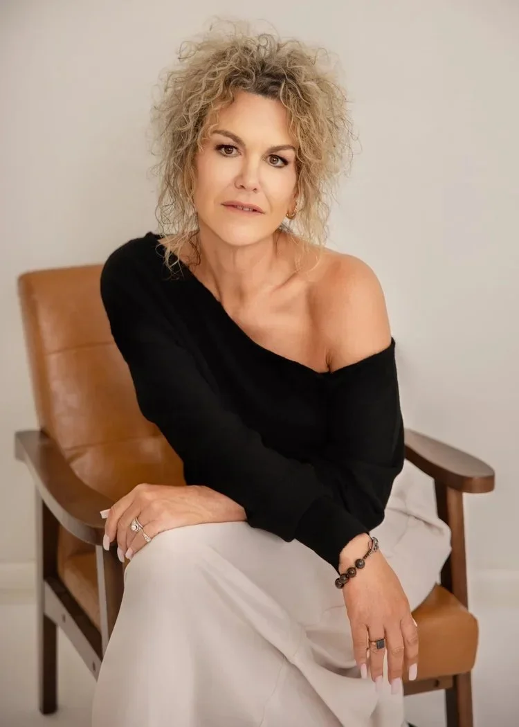 A woman with curly blonde hair sitting on a wooden chair, wearing a black off-the-shoulder top and white pants, in a neutral room.