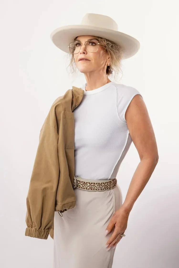 A woman with curly hair wearing a wide-brimmed white hat, glasses, a white sleeveless top, beige skirt, a patterned belt, and holding a tan jacket over her shoulder, standing against a plain white background.