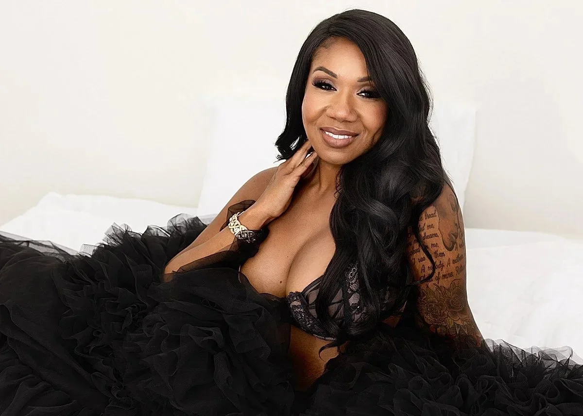 A woman with long black hair and a floral tattoo on her arm is lying on a bed, wearing black lace lingerie and a black ruffled skirt, with a relaxed and smiling expression.