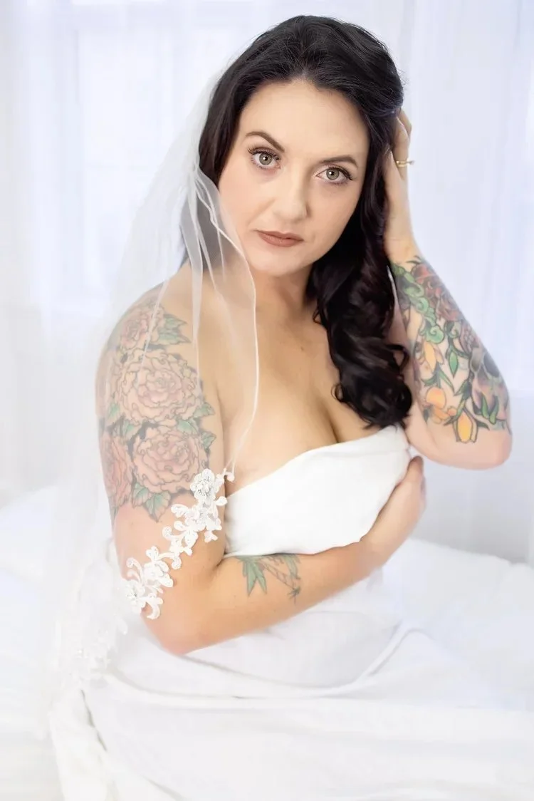 A woman with dark hair, green eyes, and tattoos on her arms is wearing a white veil and a strapless white dress. She is holding a white cloth and looking into the camera with a neutral expression.
