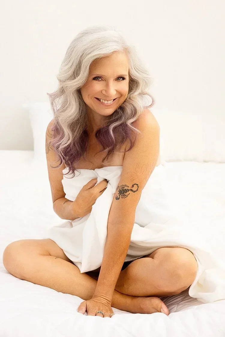 A smiling older woman with silver hair and purple tips sitting on a bed with white sheets, holding a white blanket, showing a tribal tattoo on her left arm.