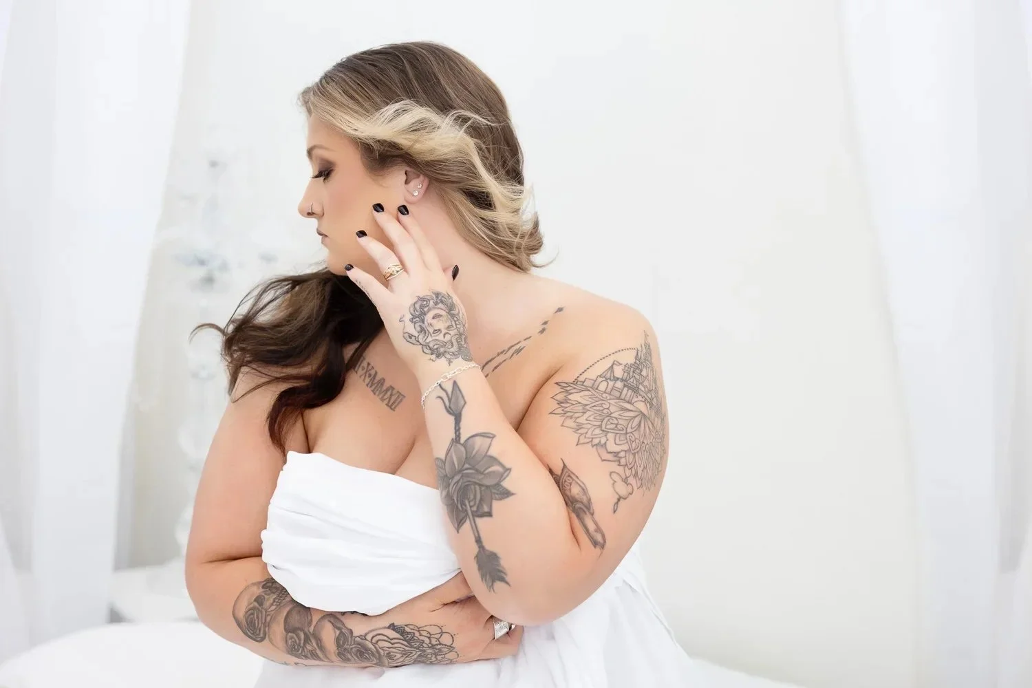 A woman with tattoos on her arms and shoulder, wrapped in a white towel, looking thoughtfully to the side in a bright, minimalist room.
