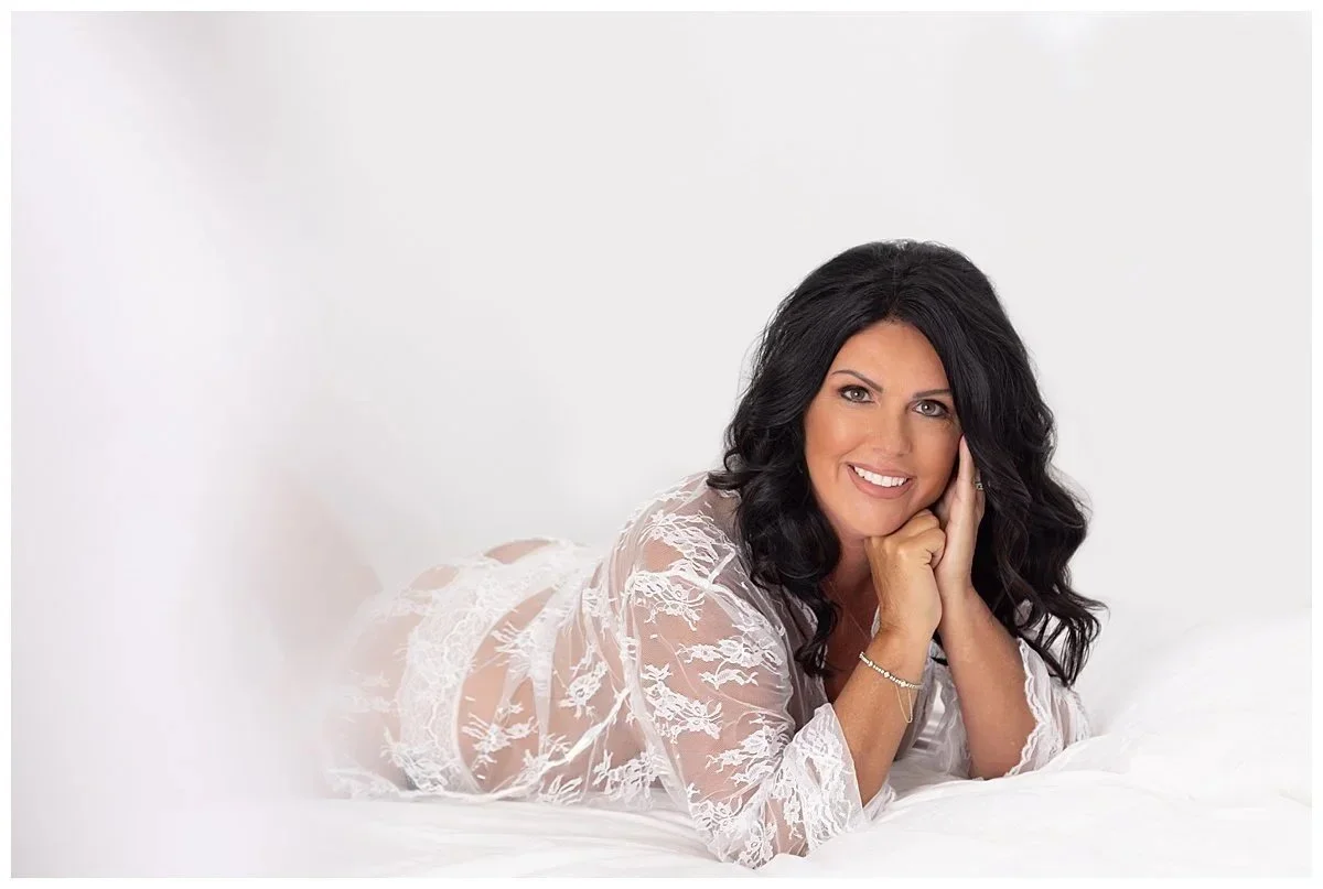 Woman with dark hair smiling, lying on a bed, wearing white lace lingerie, in a bright, white room.