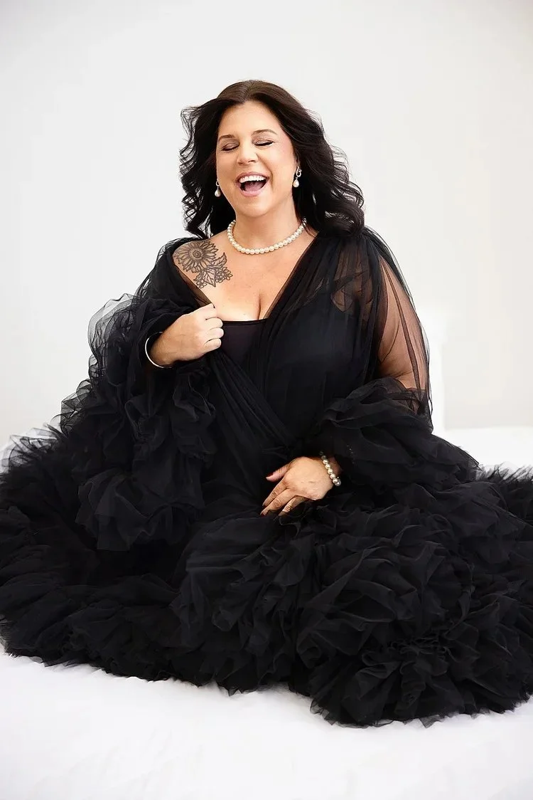 Woman in a black ruffled gown, pearl necklace and earrings, wearing a bracelet, with dark curly hair, sitting on a white surface against a plain white wall, smiling and laughing.
