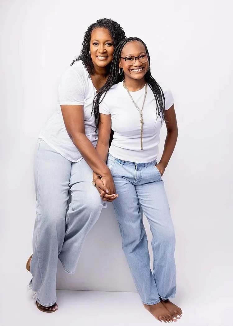 Two women smiling, holding hands, and standing against a plain white background. Both are wearing white t-shirts and light blue jeans.