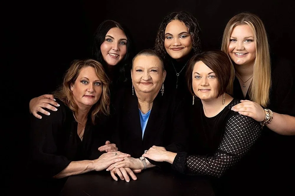 Generational family of  women posing together against a black background, smiling and embracing each other.