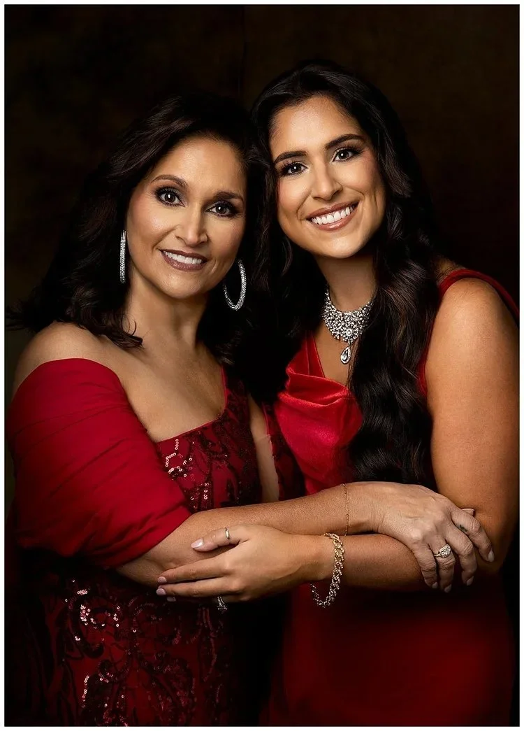 Mother and daughter  dressed elegantly in red gowns, smiling and embracing each other in a photo, wearing jewelry including earrings, necklaces, and rings.