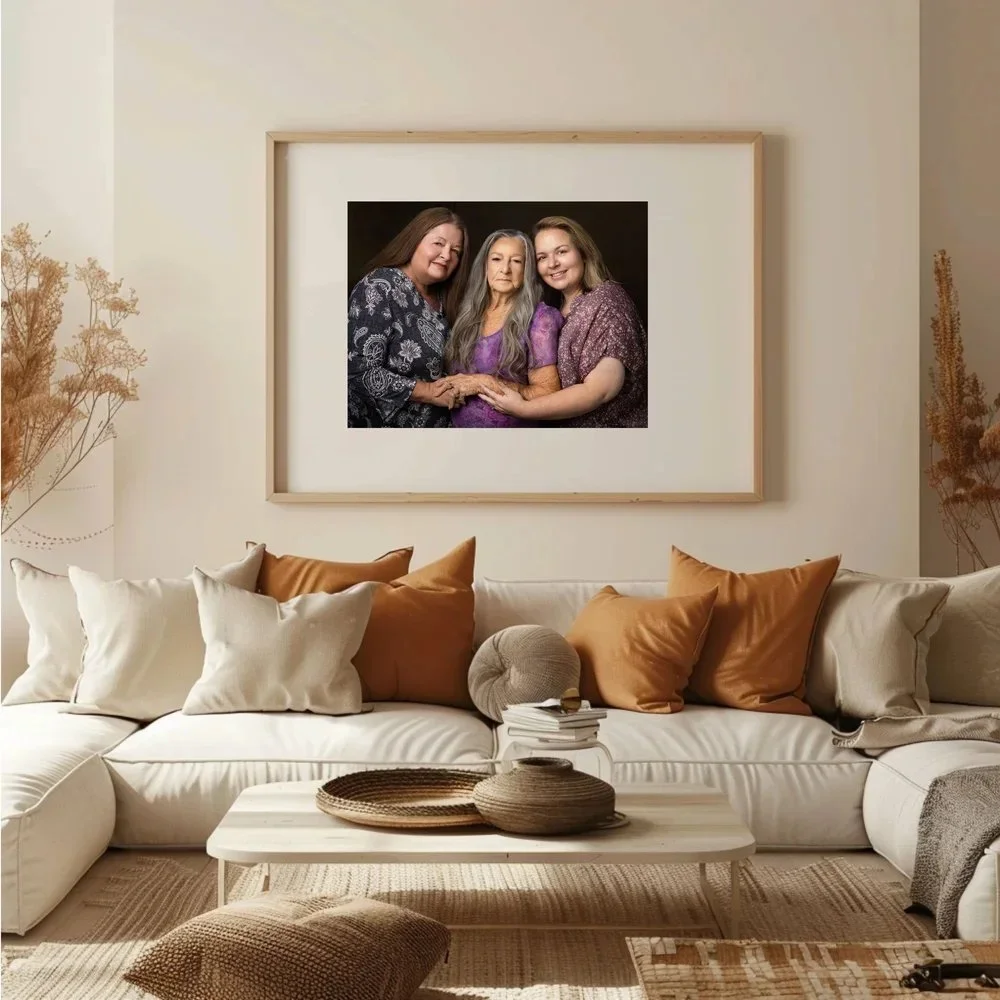 A living room with a large white sectional sofa decorated with white, beige, and rust-colored pillows. A white wooden coffee table in front of the sofa has woven baskets and a stack of books. Above the sofa hangs a framed photograph of three women wi