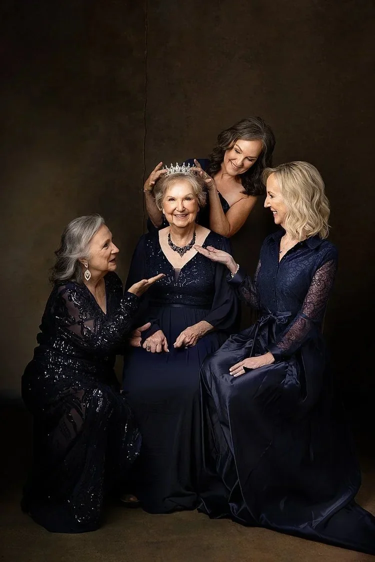 Mother with her 3 daughter's  dressed in formal dark gowns, with one woman wearing a tiara and being crowned, surrounded by three others smiling and interacting with her, in a celebratory moment.
