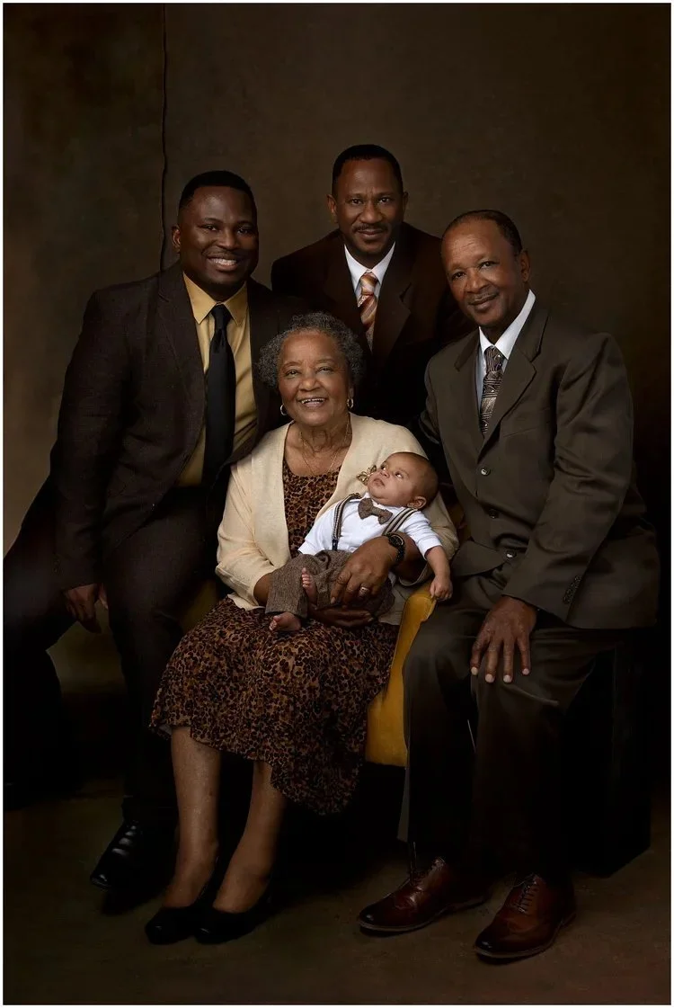 Family portrait of an elderly woman holding a baby, with four men standing around her, all dressed in formal attire, against a dark background.