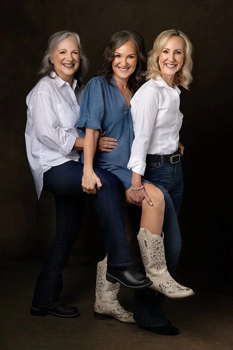 Three sisters  smiling, holding each other, against  a dark background. 