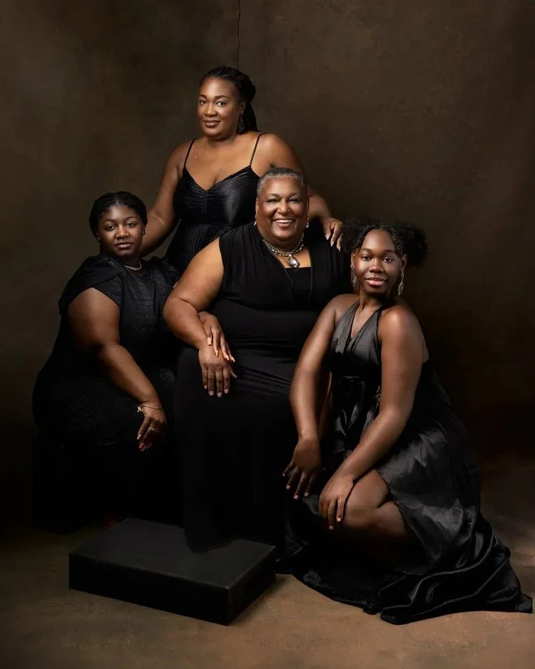 3 generations of family  in black dresses posing in a studio with a brown background, one seated, two standing, and one kneeling.