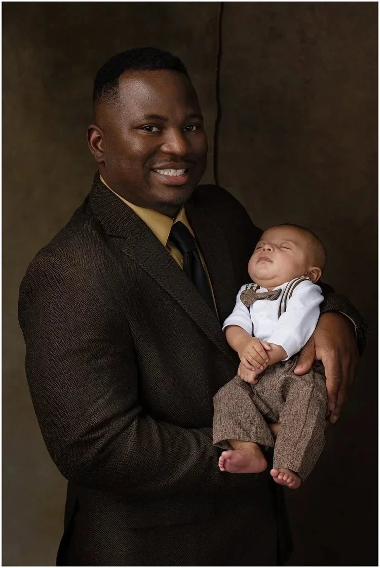 A smiling dad in a dark suit holding a sleeping baby dressed in a white shirt with a bow tie and brown pants.