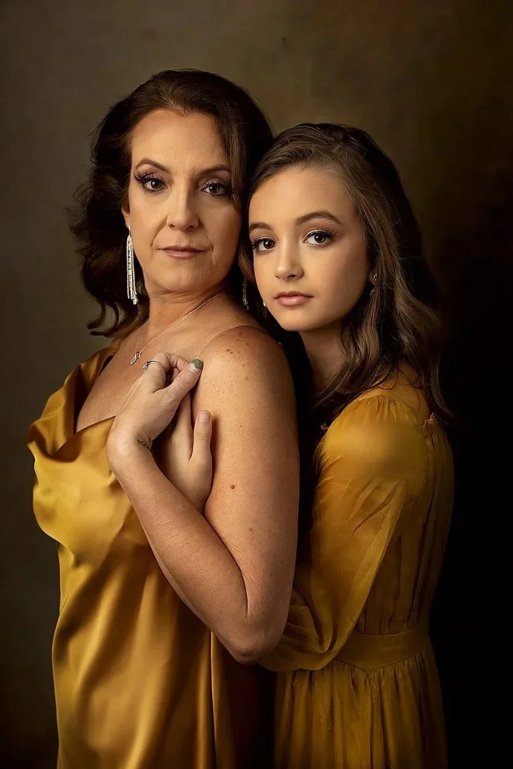 A middle-aged mom and her daughter wearing matching gold dresses posing together against a dark background.