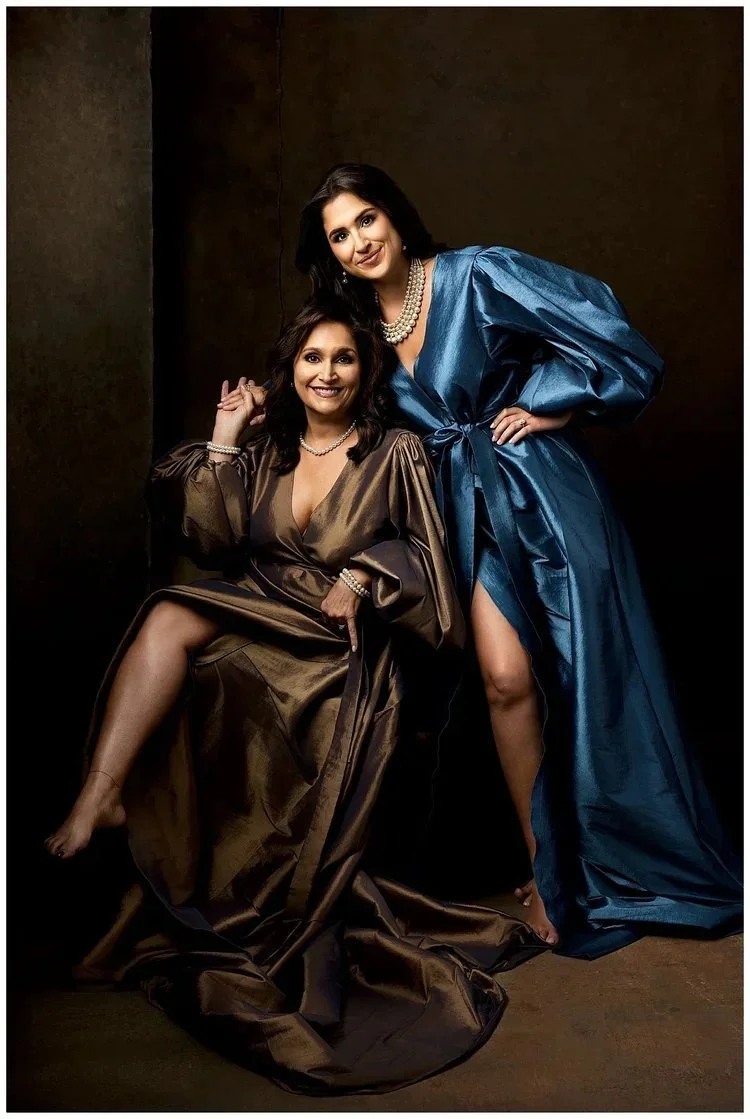 Mother and daughter  in evening gowns posing together against a dark background. One is standing in a blue gown with puffed sleeves and a slit, and the other is sitting in a brown gown with long sleeves. Both are smiling and wearing pearl necklaces.