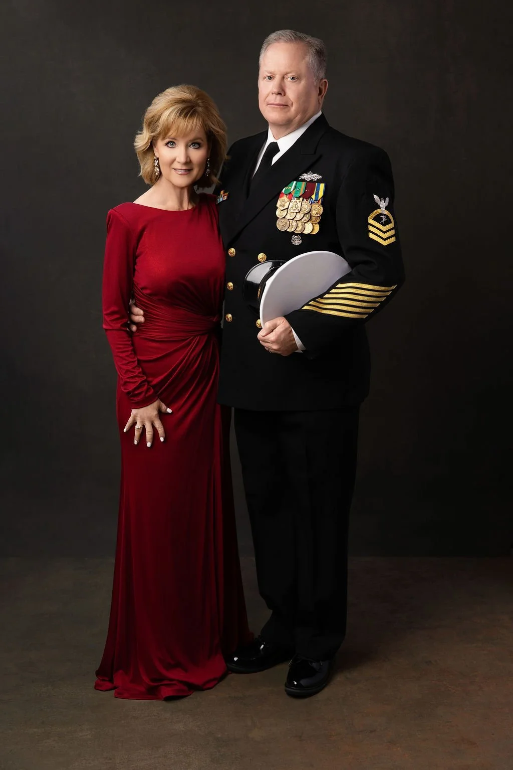 A woman in a red gown standing next to a man in a military uniform with medals, holding a white hat, against a plain dark background.