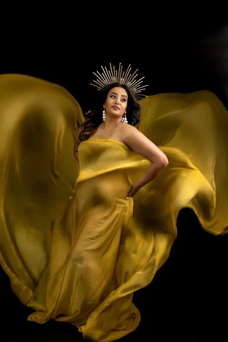 A pregnant woman dressed in a flowing gold gown with her hand on her hip, wearing a crown-like headpiece and large earrings, standing against a black background.
