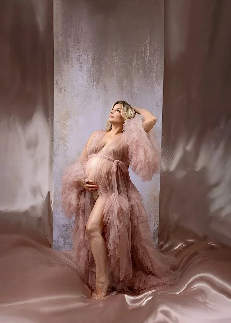Pregnant woman in a flowing, blush pink tulle dress, standing against a textured backdrop, posing with one hand on her belly and the other behind her head.