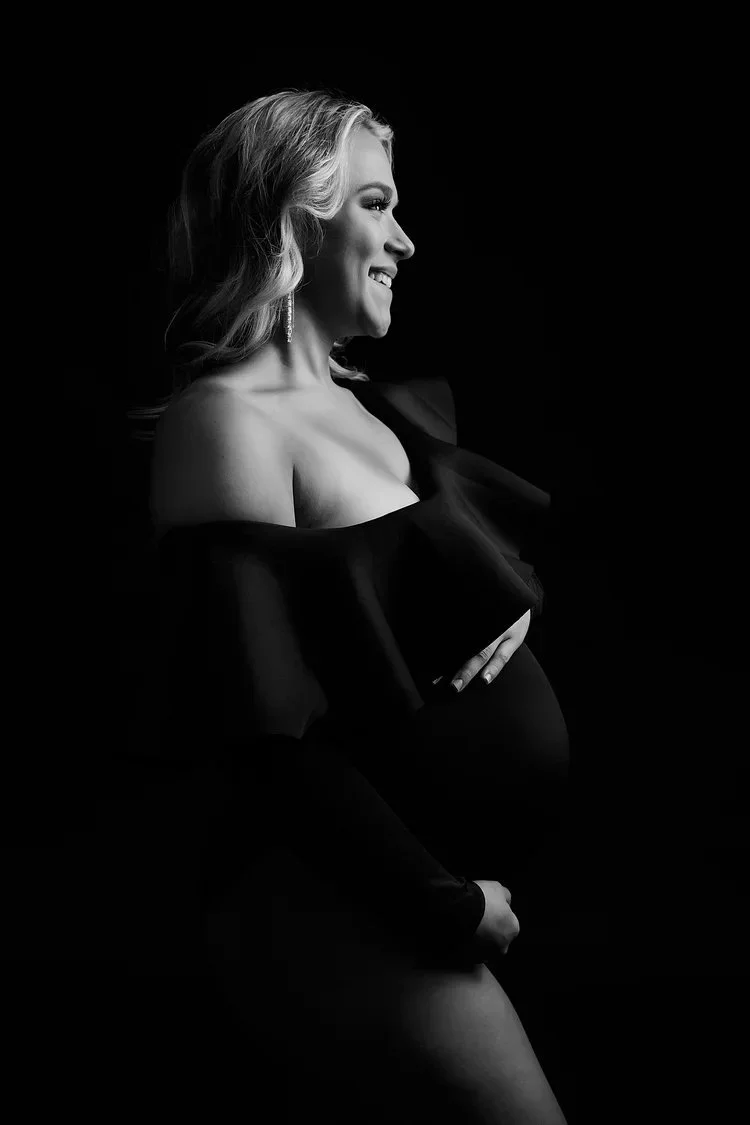 Black and white portrait of a pregnant woman with wavy hair, smiling, wearing an off-shoulder dress and earrings, against a dark background.