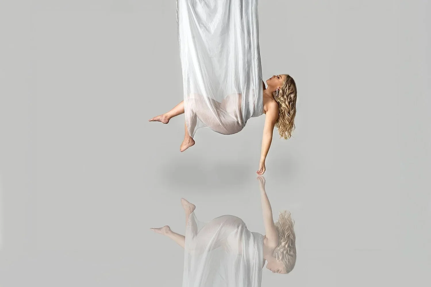 A pregnant woman with blonde curly hair in a white dress is hanging upside down with one hand touching the reflective surface, creating a mirror image of herself on the ground.