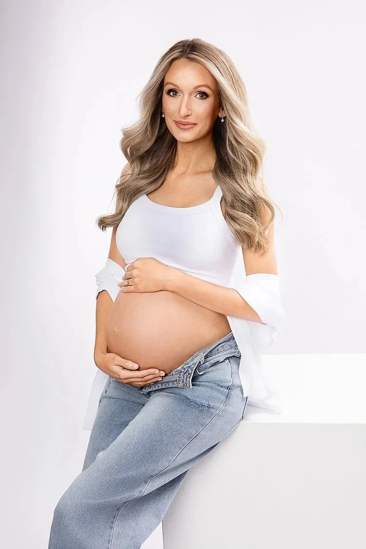 Pregnant woman with long wavy blonde hair, wearing a white tank top and light blue jeans, sitting on a white surface against a plain white background, smiling softly and gently holding her belly.
