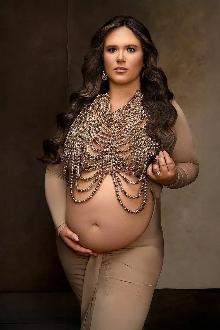 A pregnant woman with long, dark, wavy hair wearing a nude-colored outfit, draped in multiple strands of pearl jewelry covering her chest, posing against a plain background.