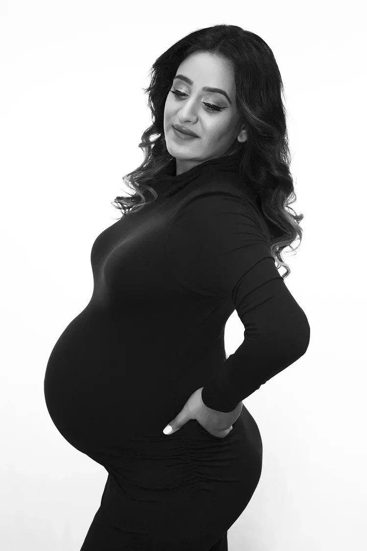 A black and white photo of a pregnant woman wearing a long-sleeve black dress, standing with her hand on her hip and looking down with a slight smile.