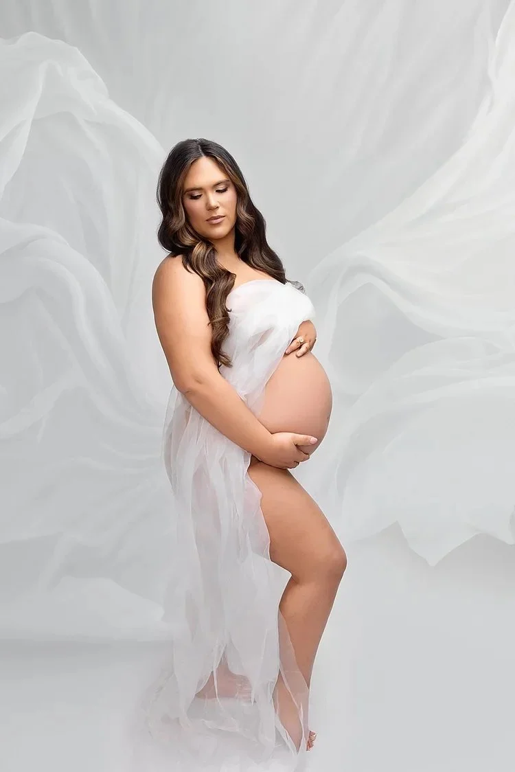 Pregnant woman with long wavy hair wearing a sheer white fabric, gently holding her belly, standing against a soft, white background.