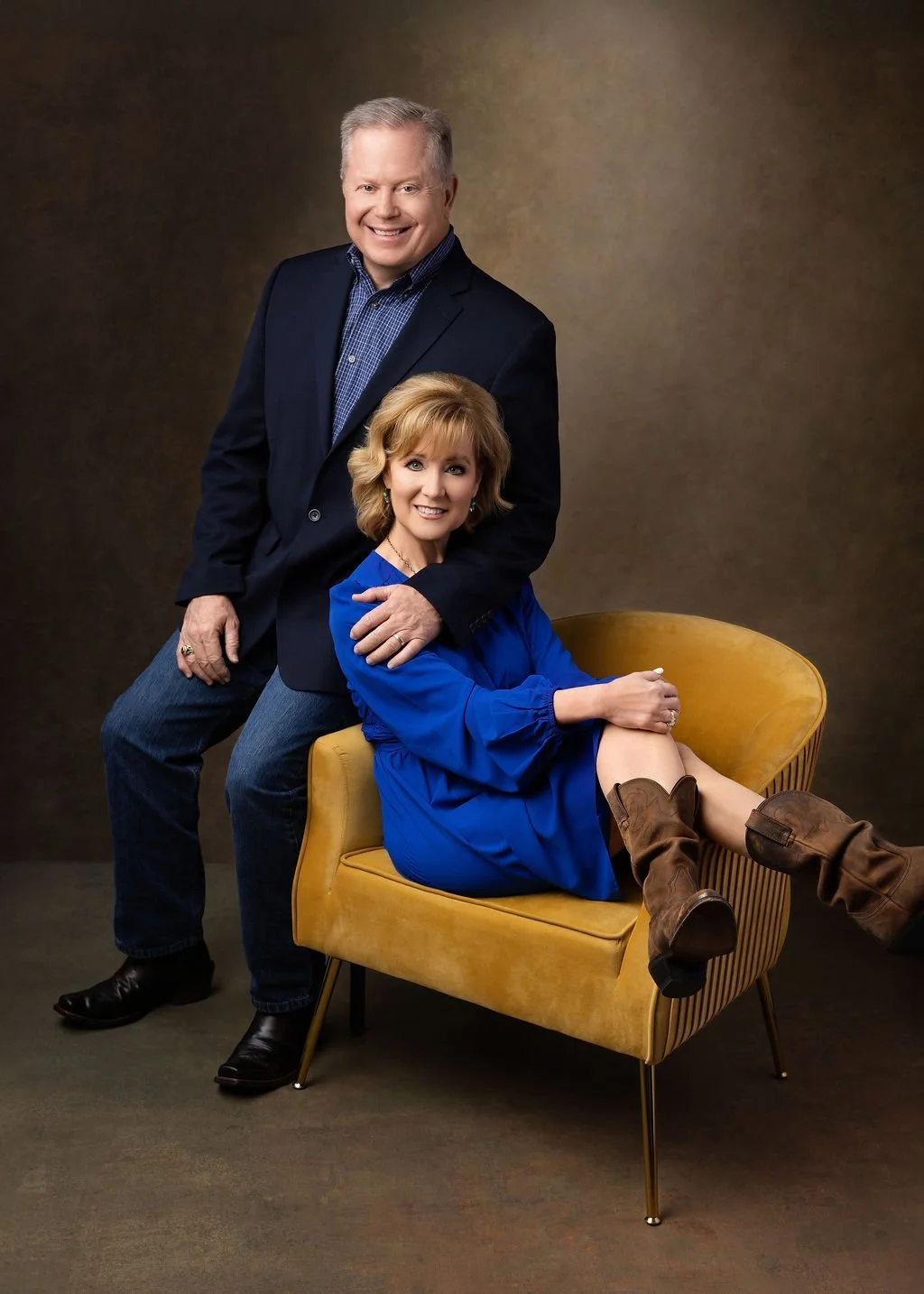 A middle-aged man and woman pose together in a studio. The woman is sitting in a mustard-colored armchair, wearing a blue dress and brown cowboy boots. The man stands beside her, resting one hand on her shoulder, wearing a dark suit jacket, blue shir