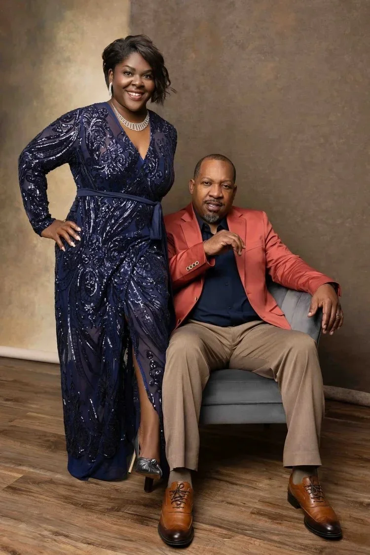 A woman in a dark blue sequined gown standing next to a seated man in a red blazer and tan pants, both smiling at the camera.