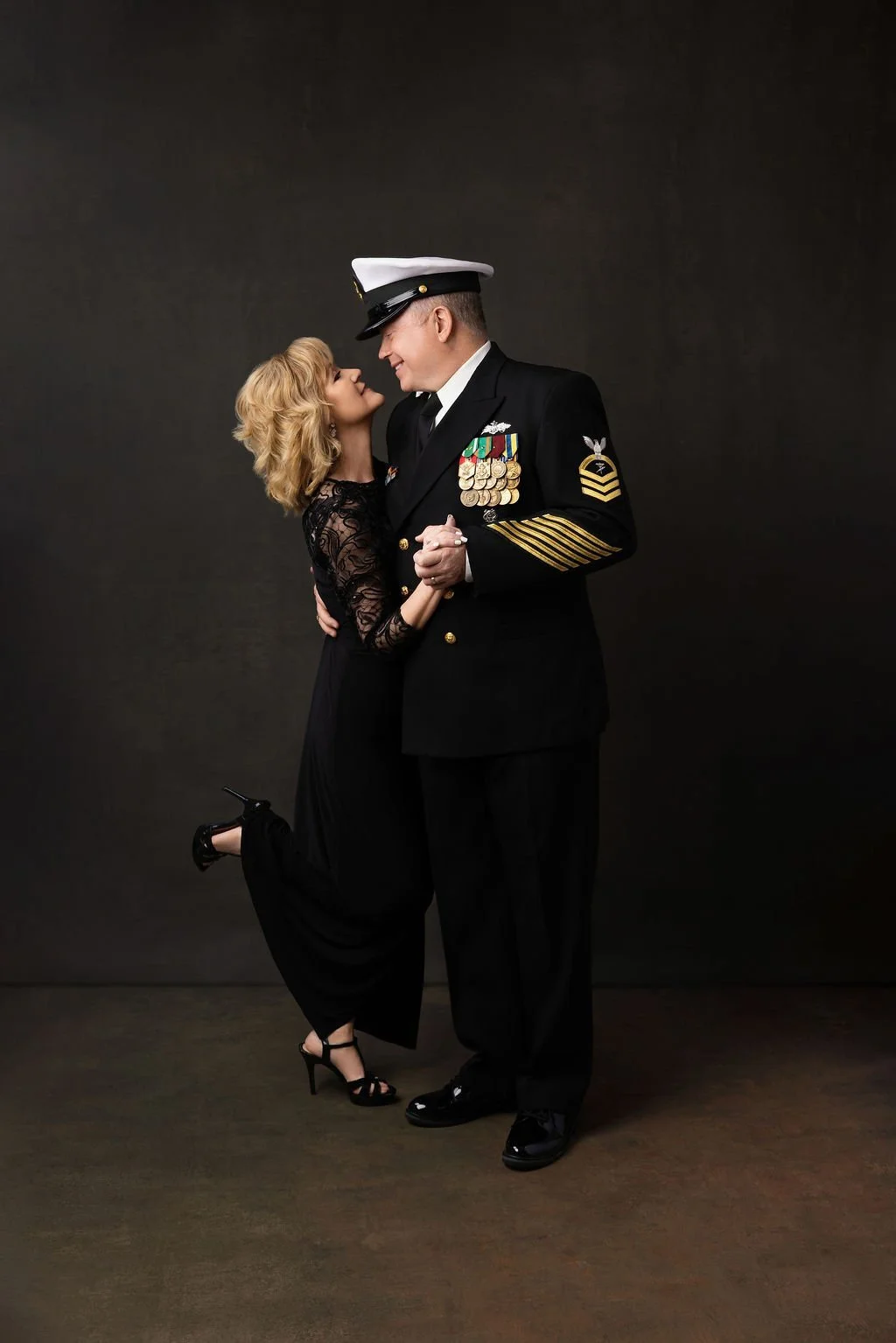 A woman in a black lace dress and heels embraces a man in a U.S. Navy uniform, who is smiling and holding her hands. They are standing close against a dark background.