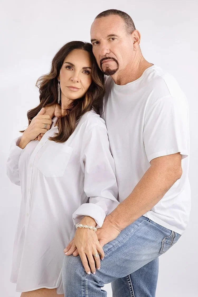 A woman and a man posing together against a plain white background. The woman has long wavy brown hair and is wearing a white shirt, earrings, and a pearl bracelet. The man has short hair, a goatee, and is dressed in a white T-shirt and jeans. They a