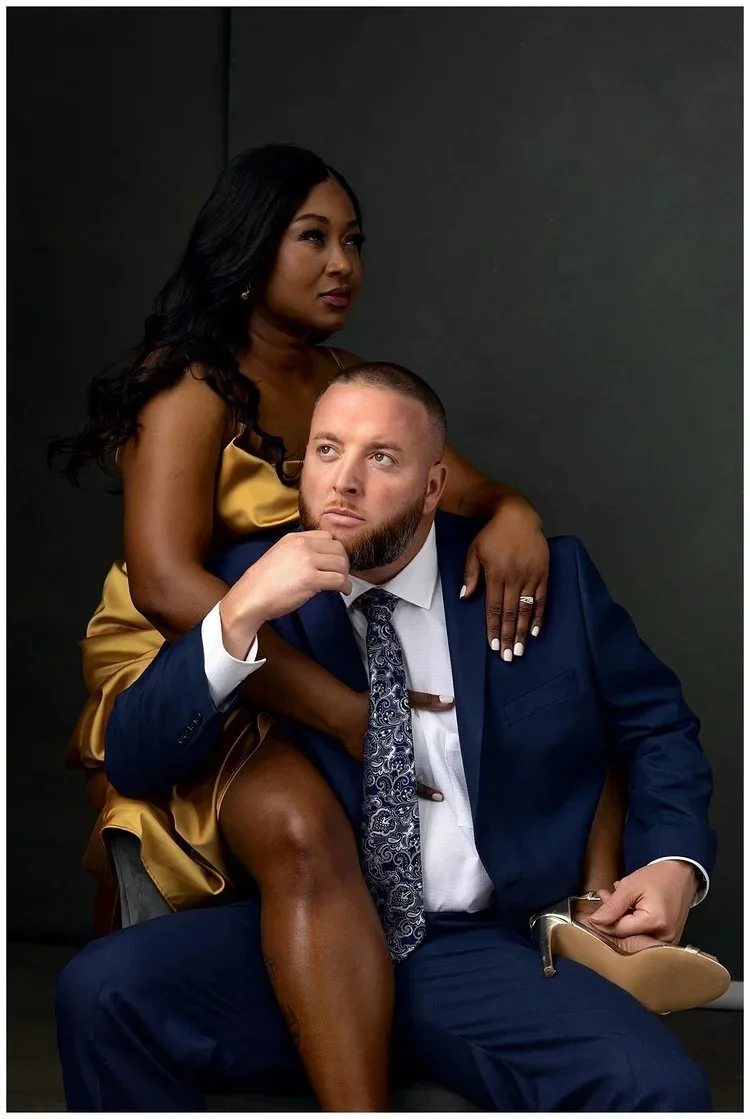 A woman in a gold satin dress sitting behind a seated man in a blue suit, with her arm around his shoulder. The man has a beard, wears a patterned tie, and holds a gold shoe in his hand. They are posed against a dark background.
