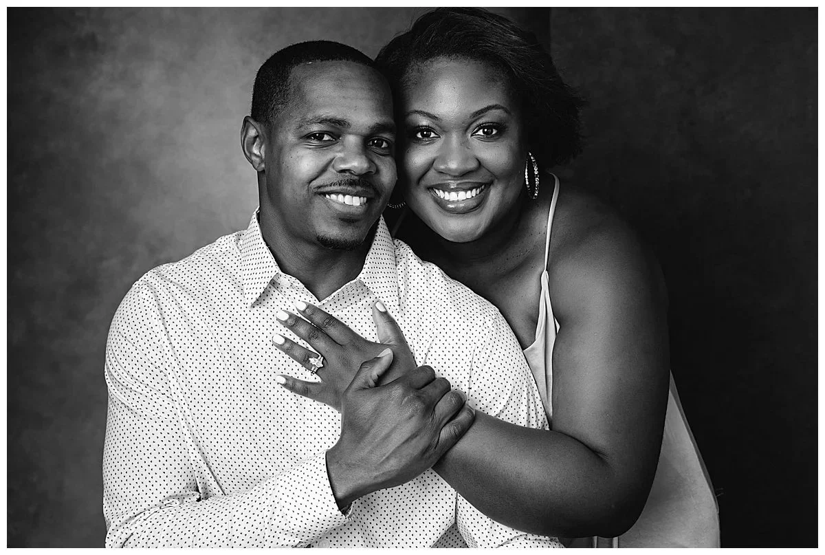 A black and white photo of a smiling man and woman posing closely together with their arms around each other. The woman is showing off an engagement ring on her finger.