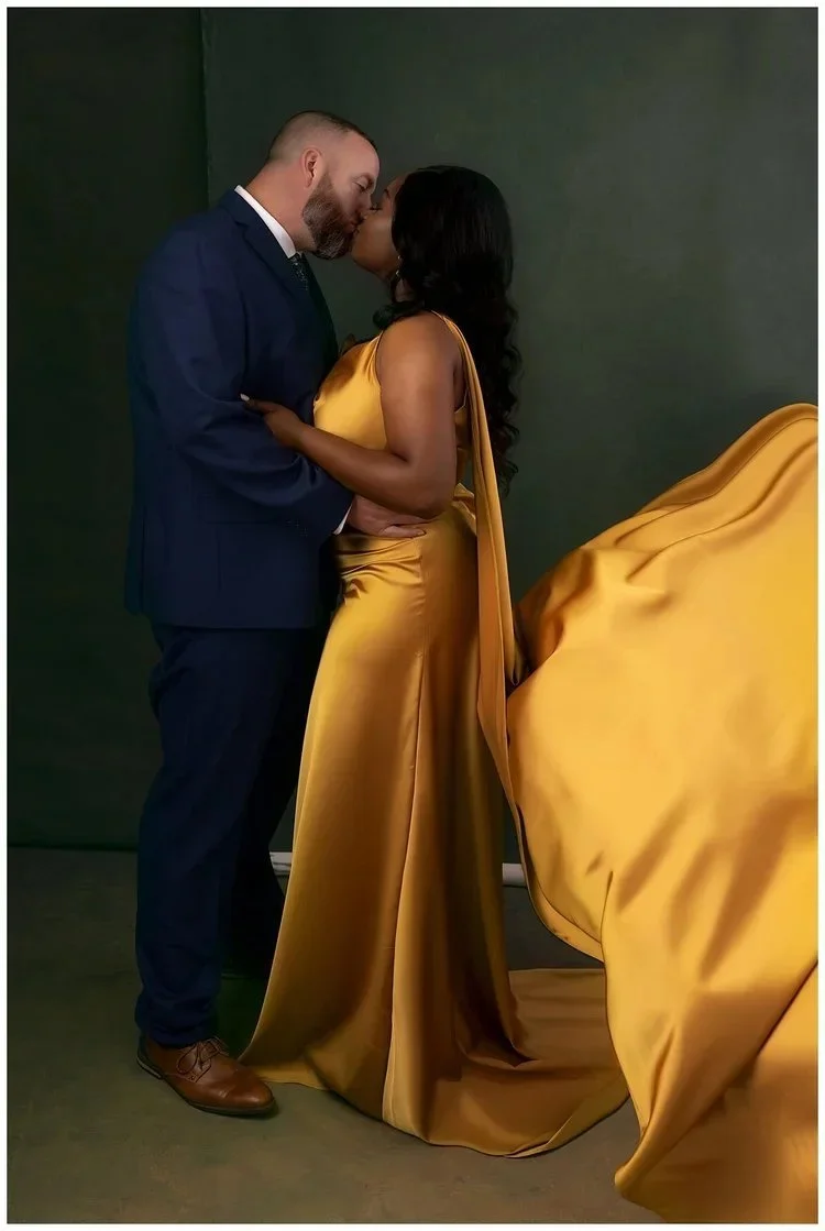 A couple in formal attire sharing a kiss, with the woman dressed in a gold satin gown and the man in a navy suit, against a dark green background.