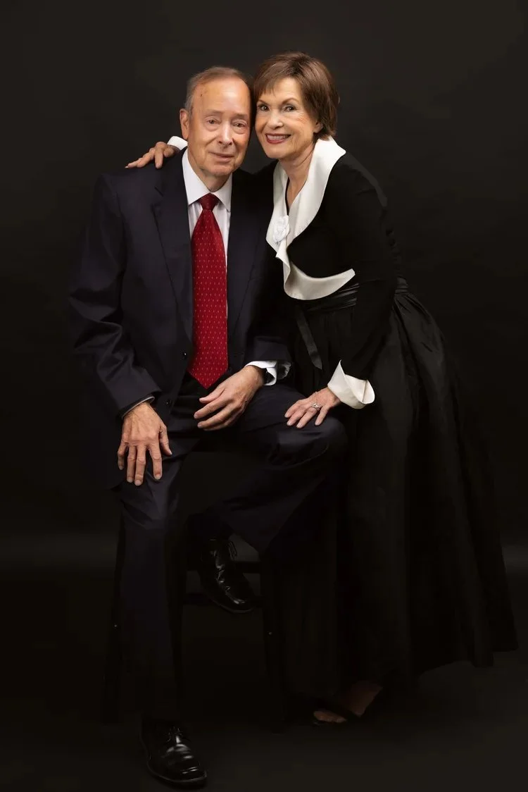 An elderly man in a suit and red tie seated on a stool next to a woman in a black dress with white accents, both smiling against a black background.