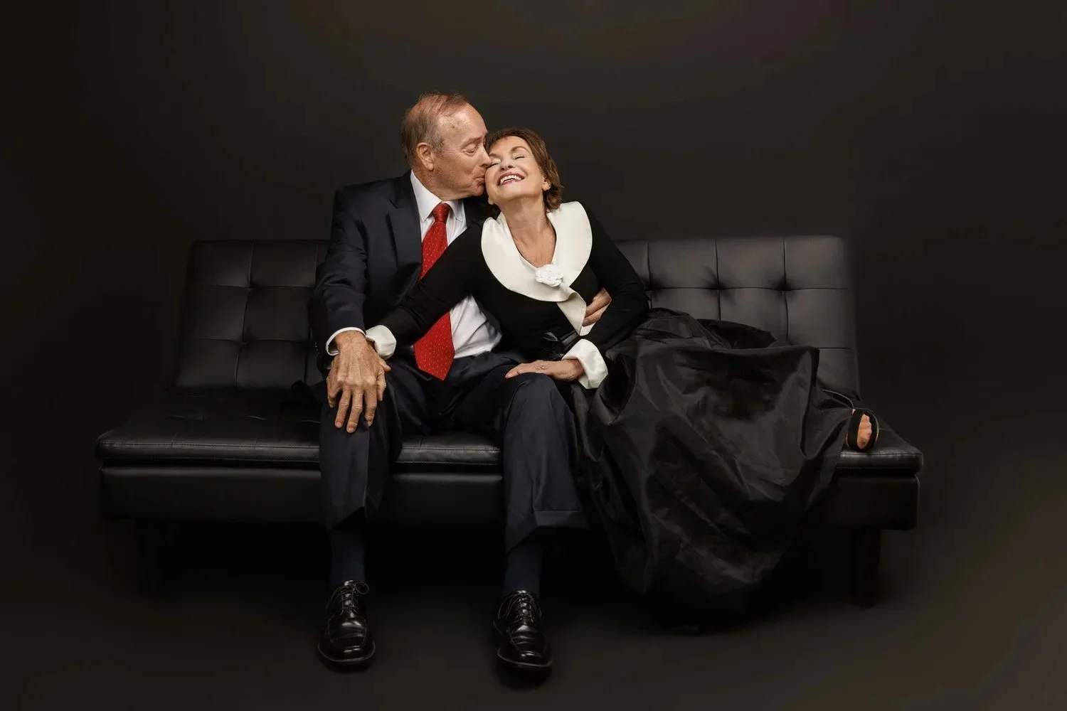 An elderly man in a suit and a woman in a black dress with white collar sitting on a black bench against a black background, sharing a tender moment with the man kissing the woman on her cheek.