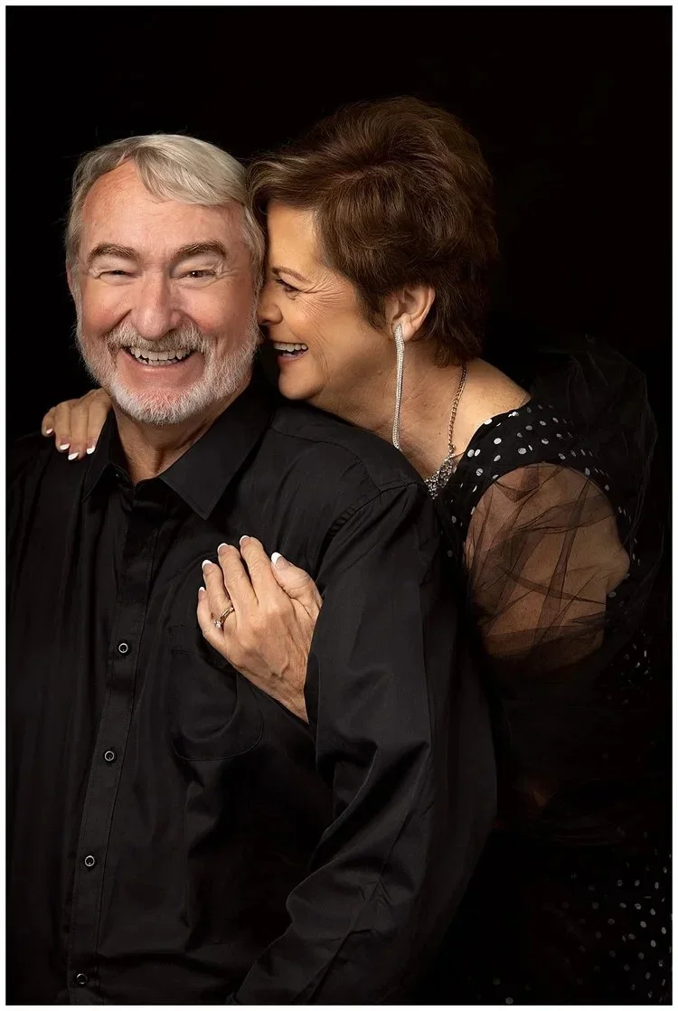 An elderly couple smiling and laughing together against a black background. The woman is leaning on the man’s shoulder with her hand on his chest, and they are both dressed in black.