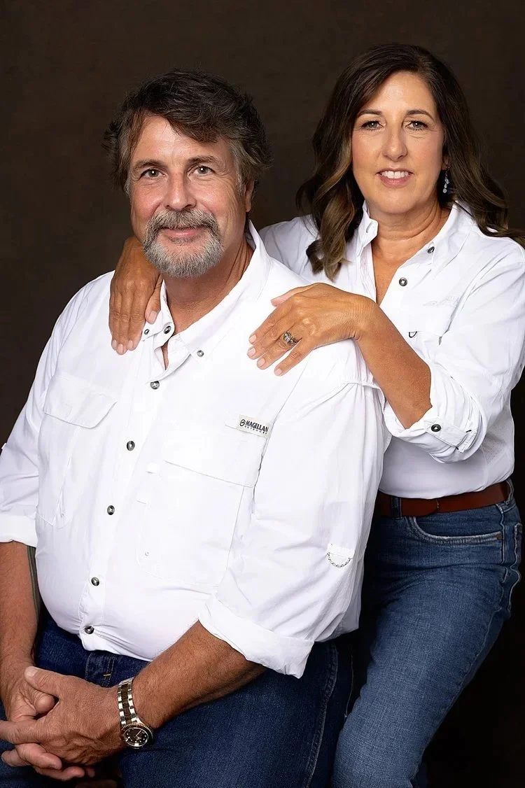 A man and woman posing together, both dressed in white shirts and jeans, with the woman resting her hand on the man's shoulder.