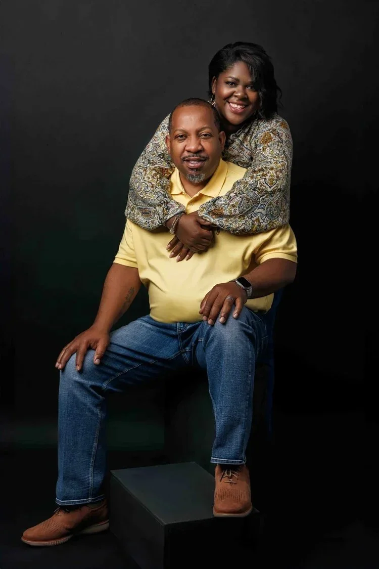 A man is sitting on a black box, wearing a yellow polo shirt and blue jeans. A woman is standing behind him, leaning over with her arms around his shoulders. She is smiling, wearing a patterned blouse, and has short curly hair. The background is blac